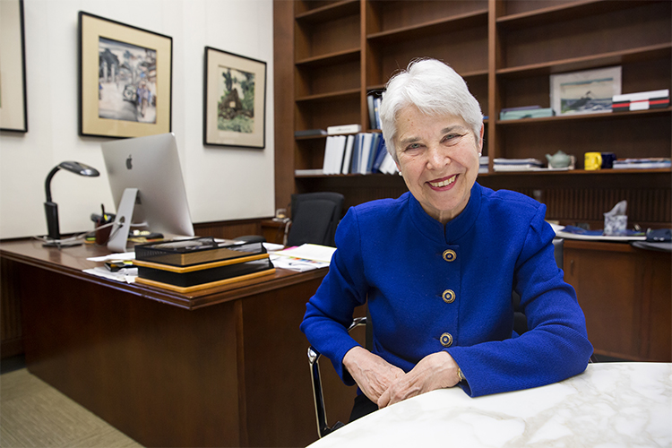 A photo of Carol Christ wearing a royal blue button up, sitting down in her office next to a brown, wooden desk while smiling and having both arms resting by her side.