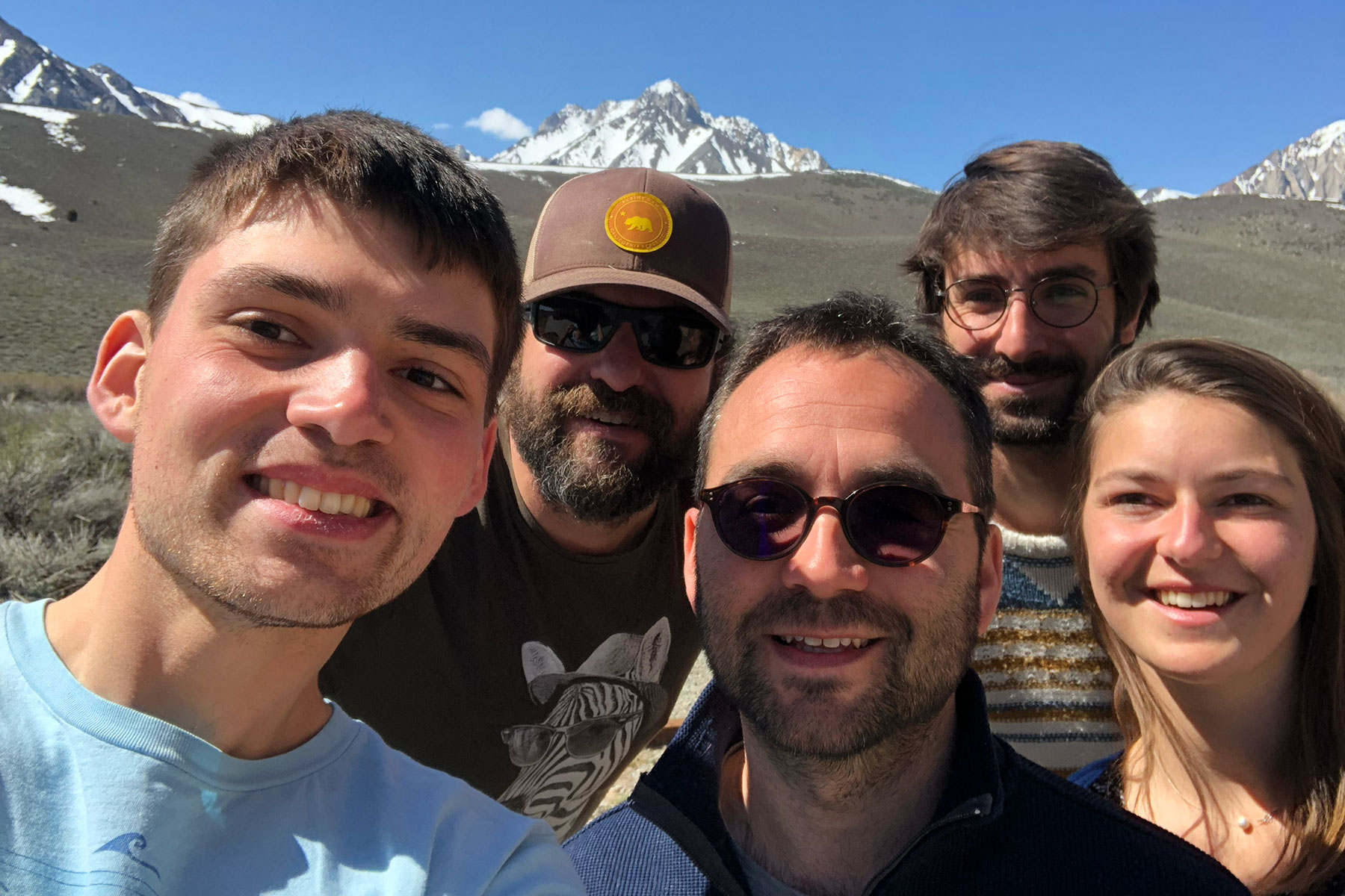Five people take a group selfie with a view of snow-capped peaks in the background.