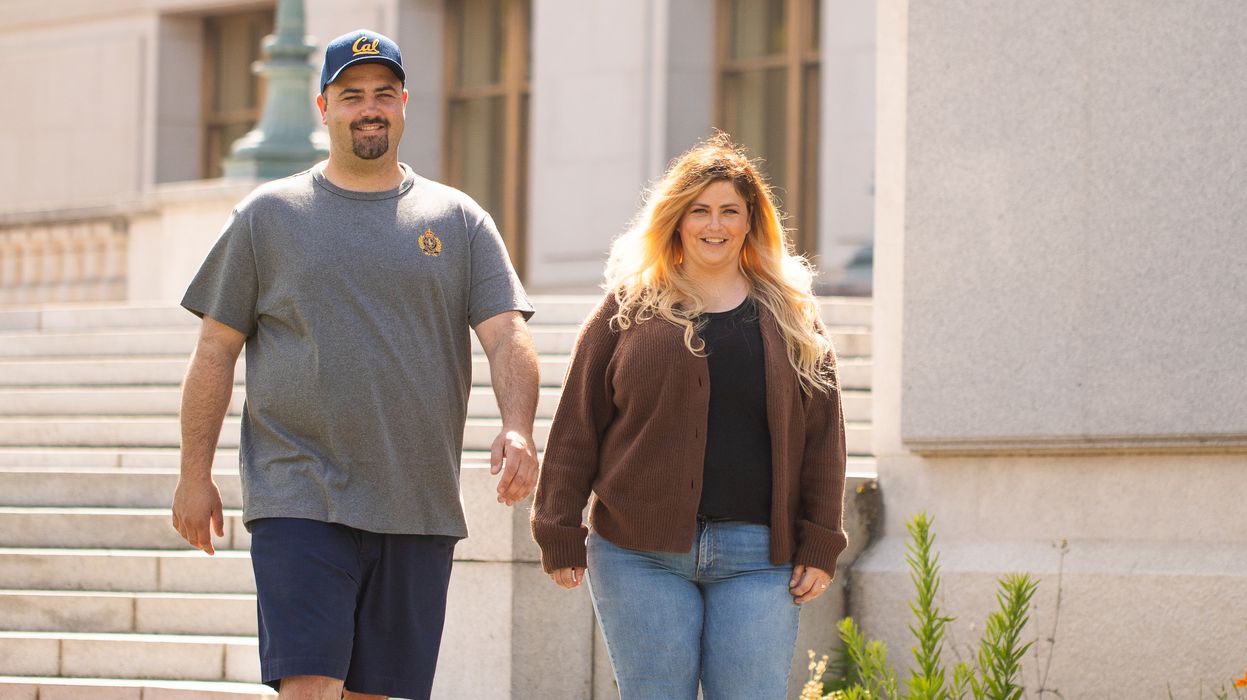 a man in a gray tshirt and woman in a brown sweater walk on a sidewalk lined with green plants outside of a concrete building