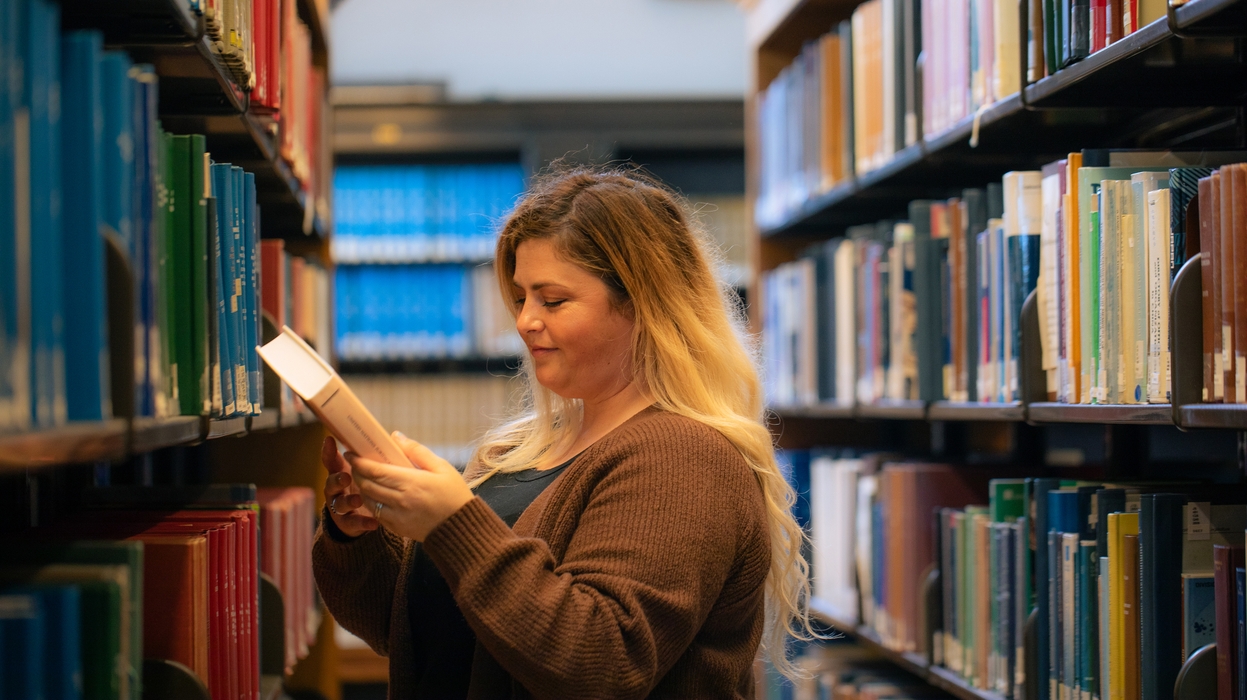 a woman looks at the cover of a book between two shelves of library books