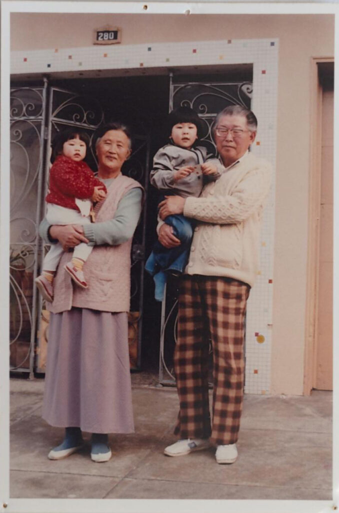 Two adults wearing light-colored jackets stand outside a building holding two young children