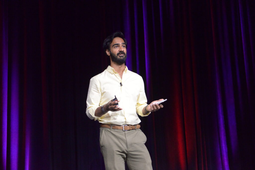 Imran Khan in a light-colored shirt and tan pants speaking on a stage during a psychedelics conference.