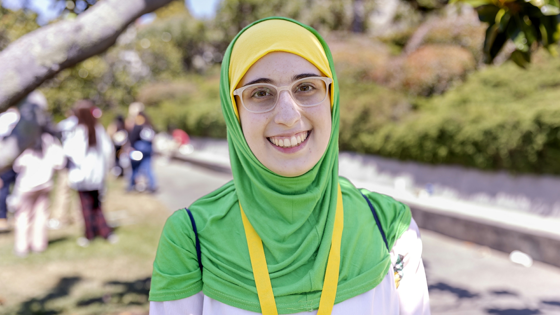 a person wearing a yellow and green headscarf and glasses stands outside and smiles