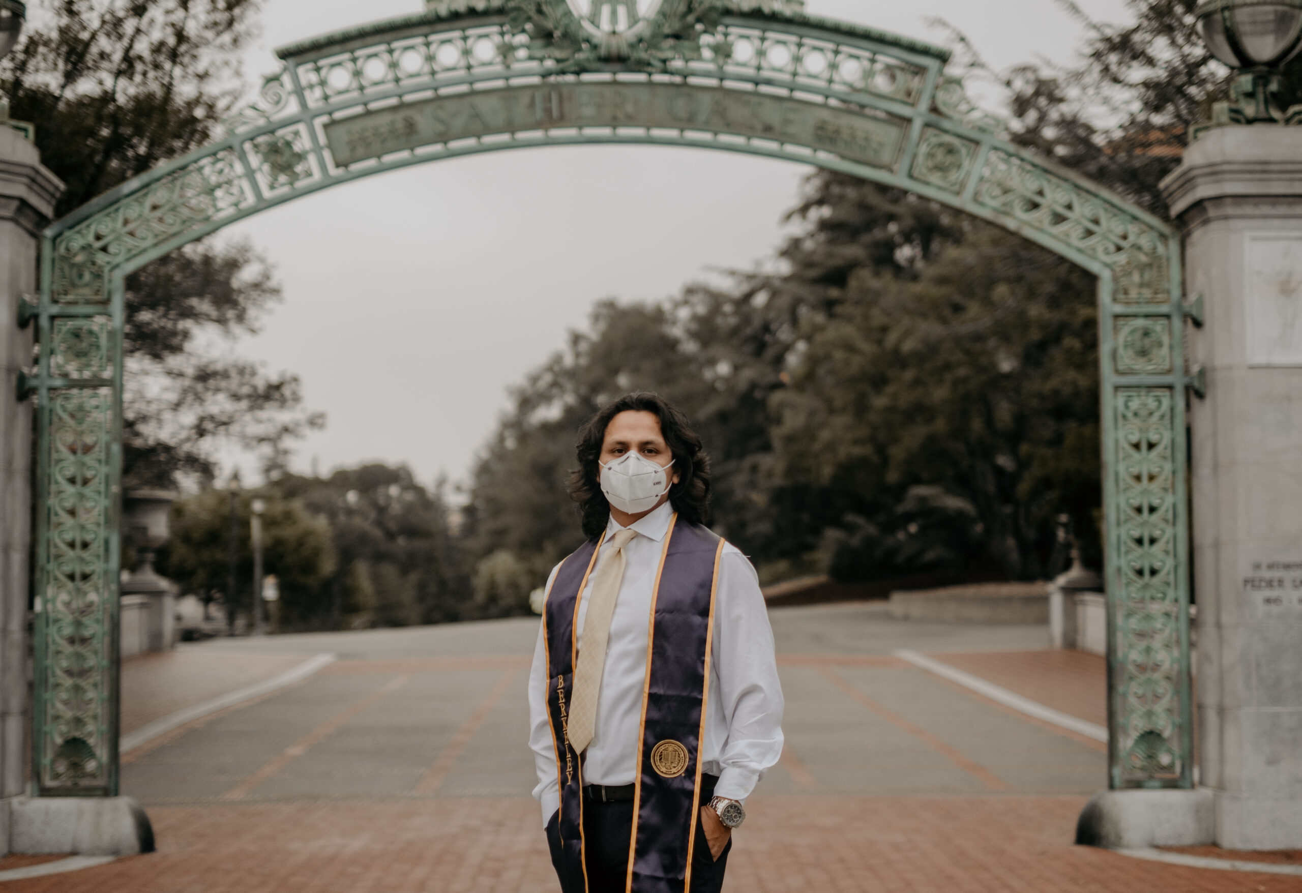 Luis posing in front of Sather Gate with a white mask, berkeley stole, and white collared shirt with black dress pants with his hands in his pockets.
