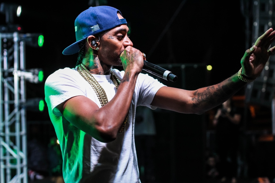 An up close photo of Nipsey Hussle wearing a blue hat and white t-shirt with a microphone to his mouth as he has both arms up.