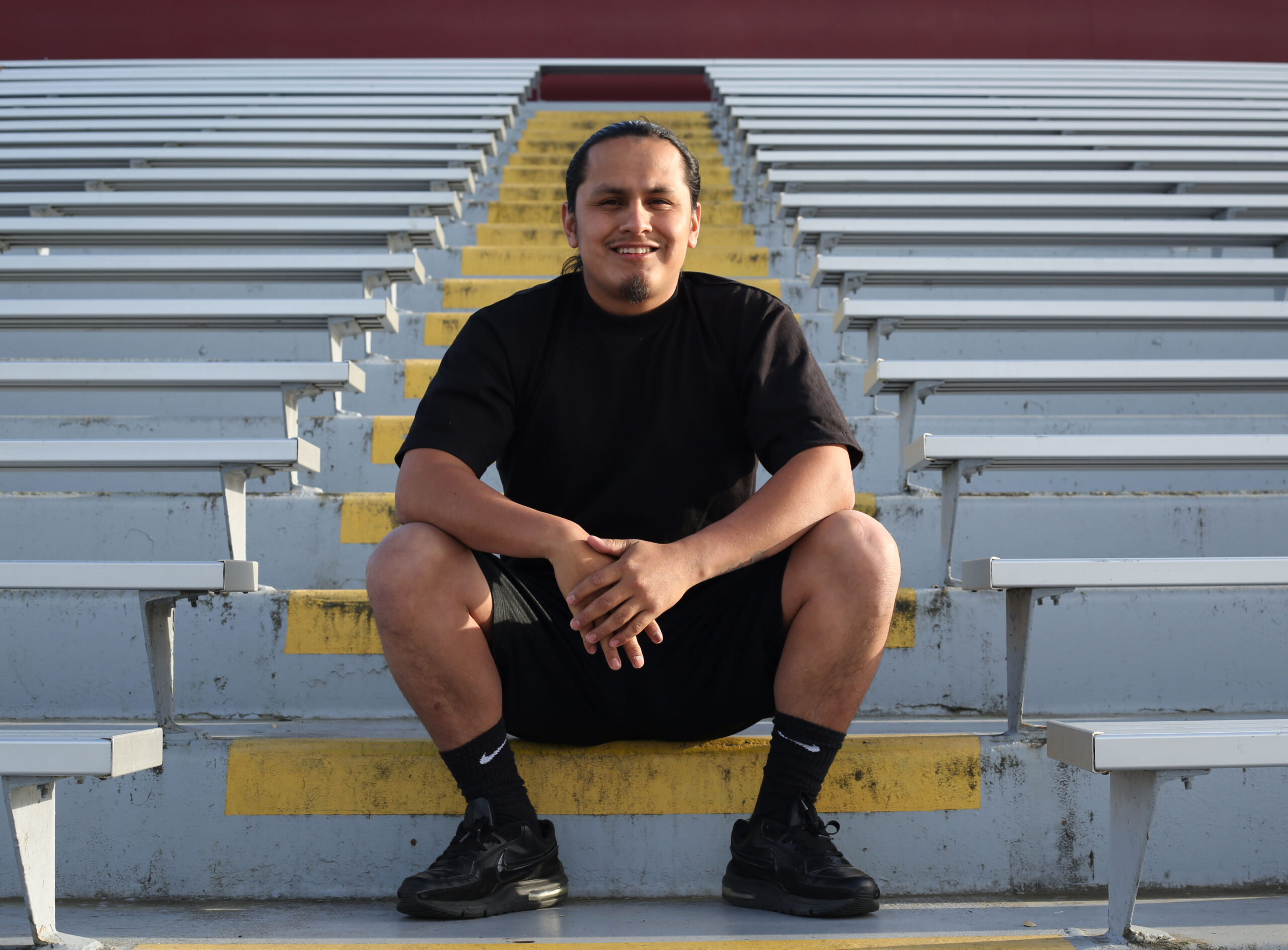 Luis sits on one of many rows of stairs behind him, wearing a black shirt, shorts, socks, and shoes as he smiles on a sunny evening.