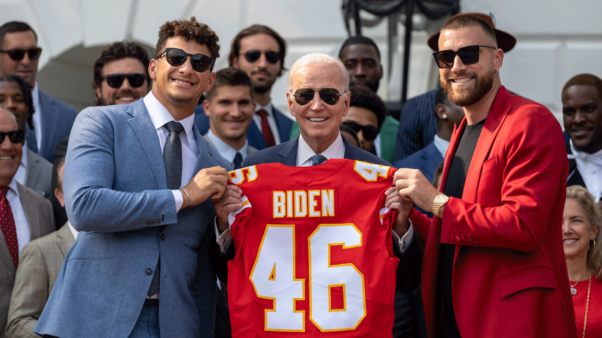 Kansas City Chiefs stars Patrick Mahomes and Travis Kelce stand on both sides of President Biden. All three are holding a red chiefs jersey with the name Biden and #46. 