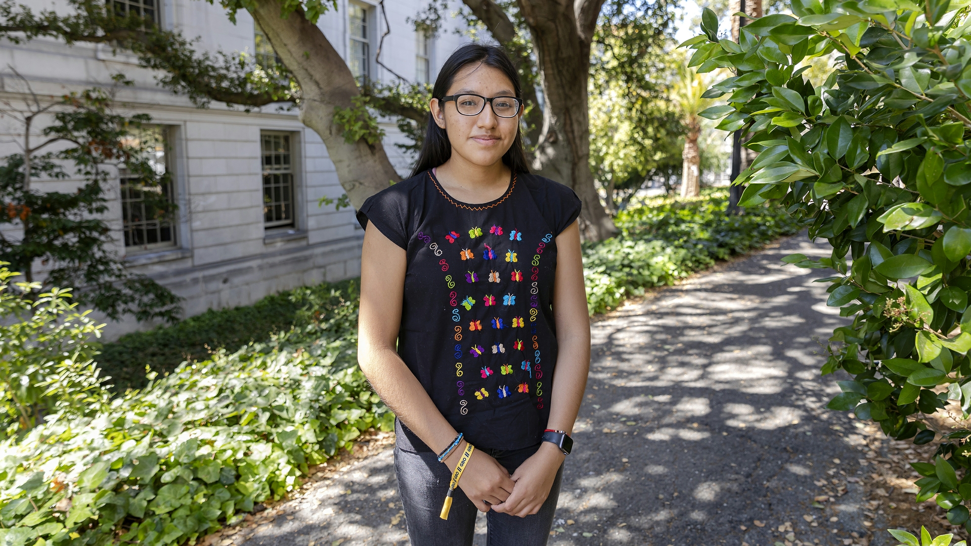 a person with long dark hair and glasses wearing a black shirt with colorful embroidered butterflies on it stands outside and smiles