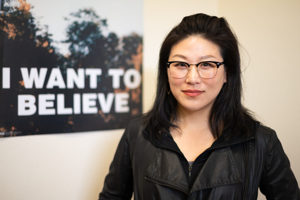 A person with long dark hair and glasses looks at a camera in front a poster with the words I Want To Believe