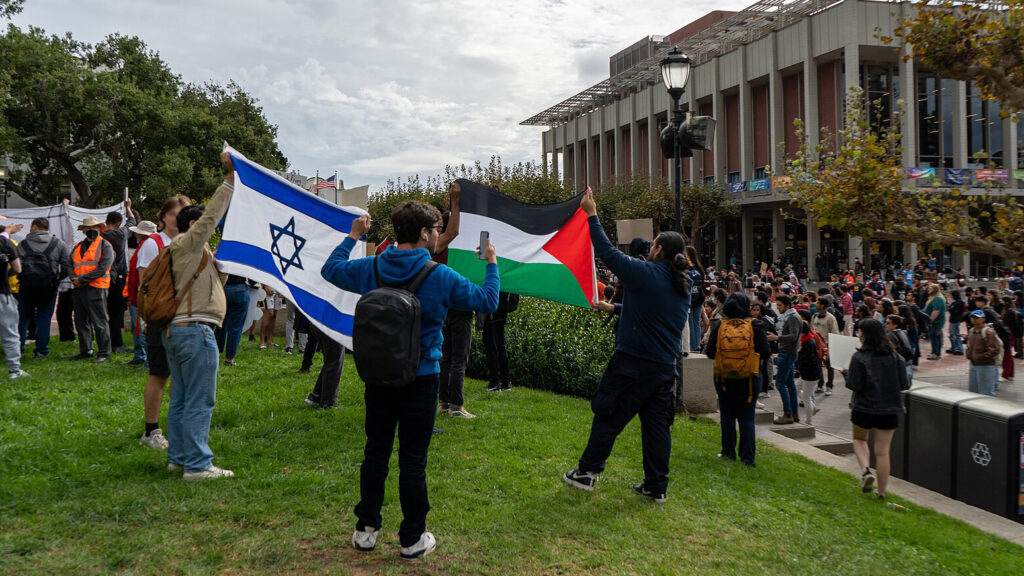 Two students hold an Israel flag and two students hold up a Palestinian flag standing next to one another on the Berkeley campus with at least one hundred students on Sproul Plaza in the background