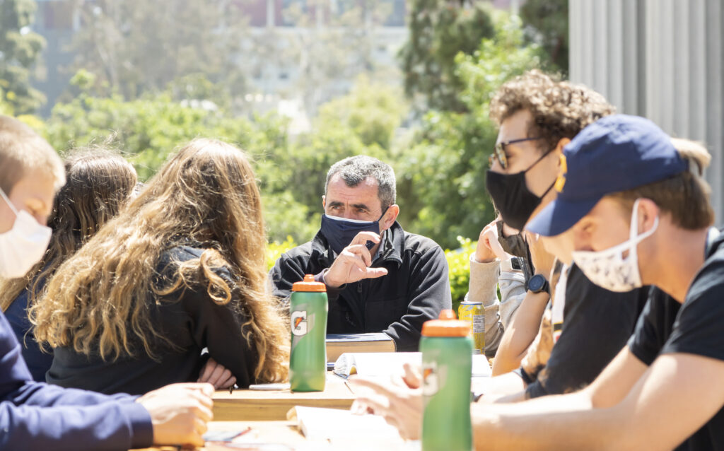 A group of students wearing face masks sitting outside looks toward a man at the end of the table gesturing with his hand