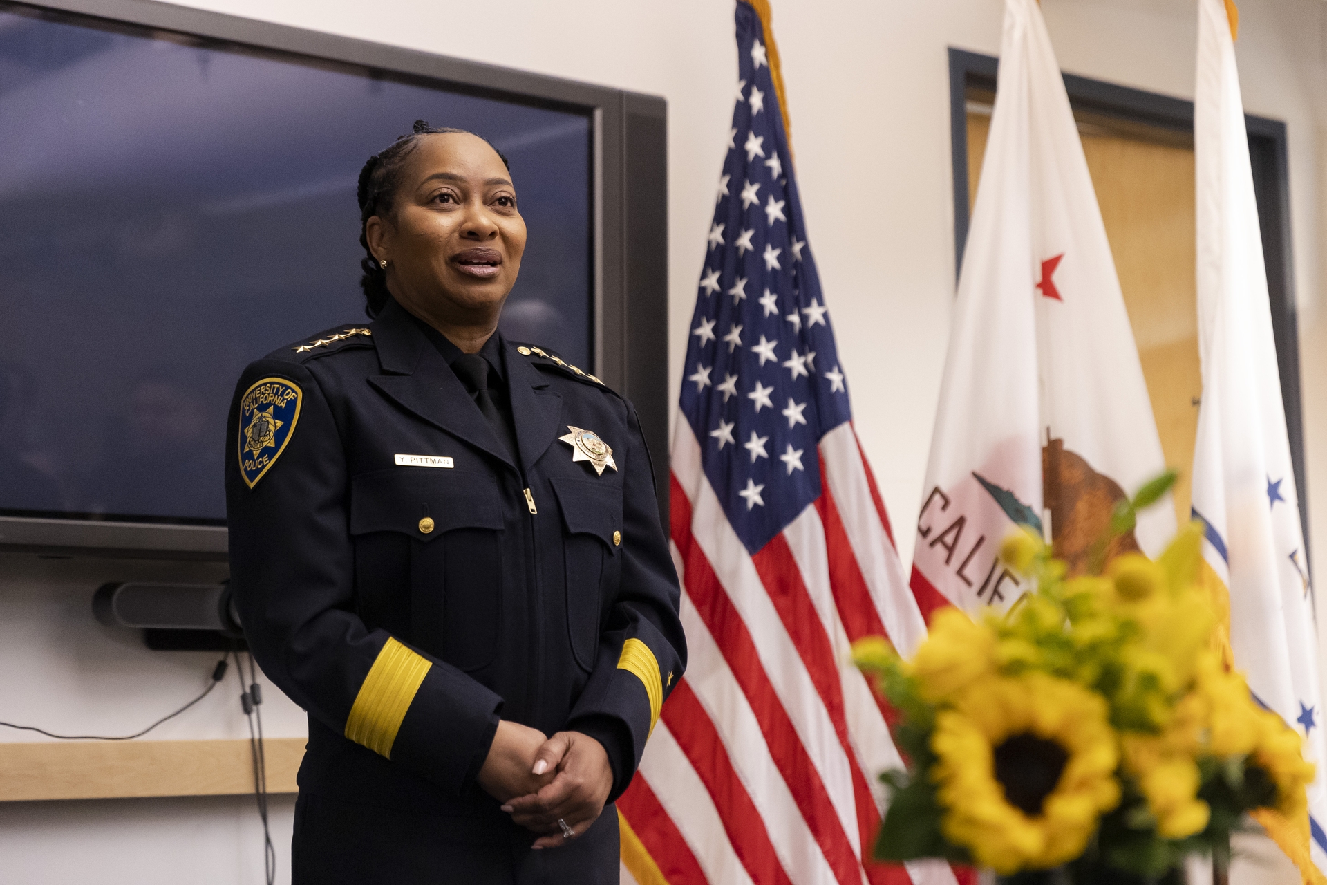 A person wearing a police chief uniform stands in front of a tv screen and U.S. and California flags