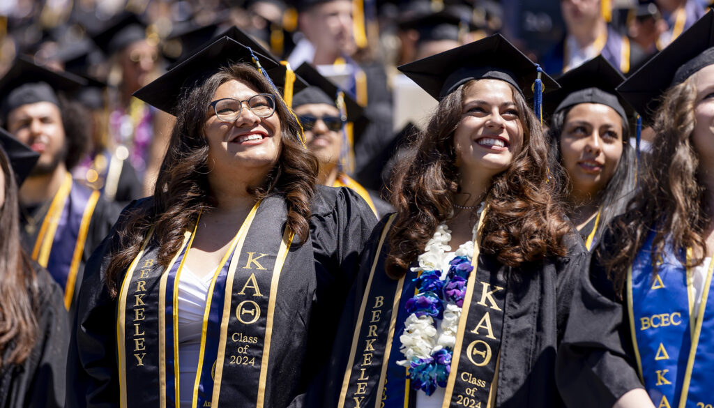 Two women wearing commencement robes, leis and stoles smiled as they celebrated.