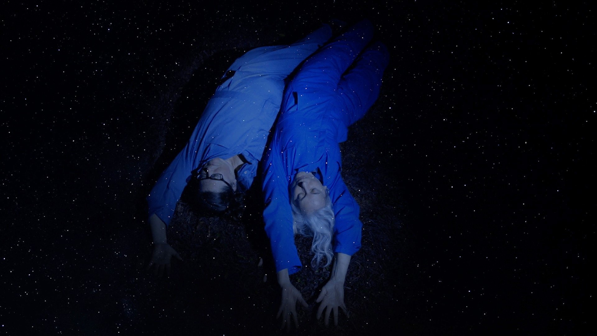 two people wearing blue jumpsuits lay back outstretched in the night sky filled with stars