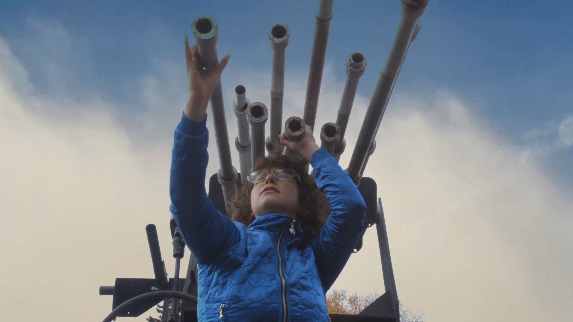 a person wearing a blue jacket stands under a set of metal pipes and holds two in their hands and looks out into the sky