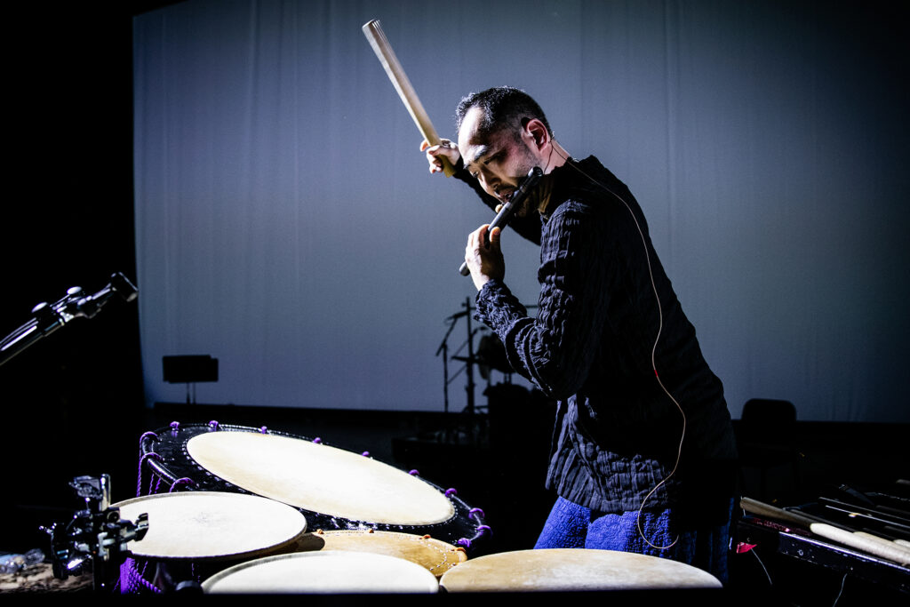 Kaoru Watanabe plays large drums and a flute at the same time