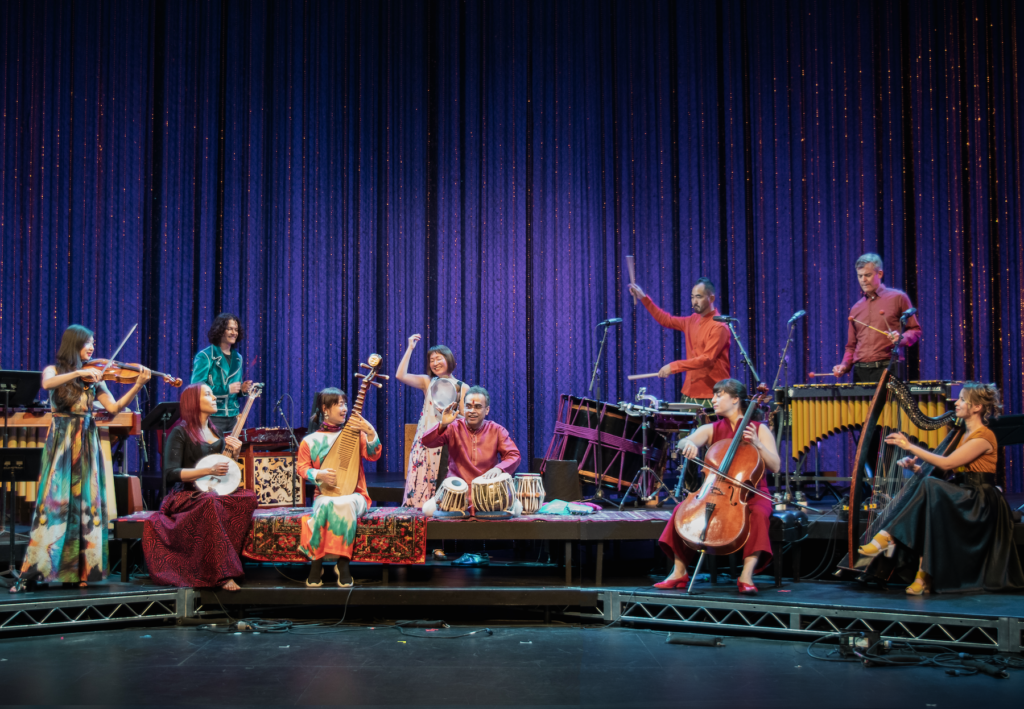 The Silkroad Ensemble of 10 musicians dressed in bright colorful clothing play instruments on stage