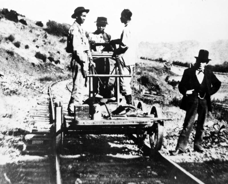a black-and-white photo of three Chinese railroad workers on a handcar with a white man in a top hat standing next to them