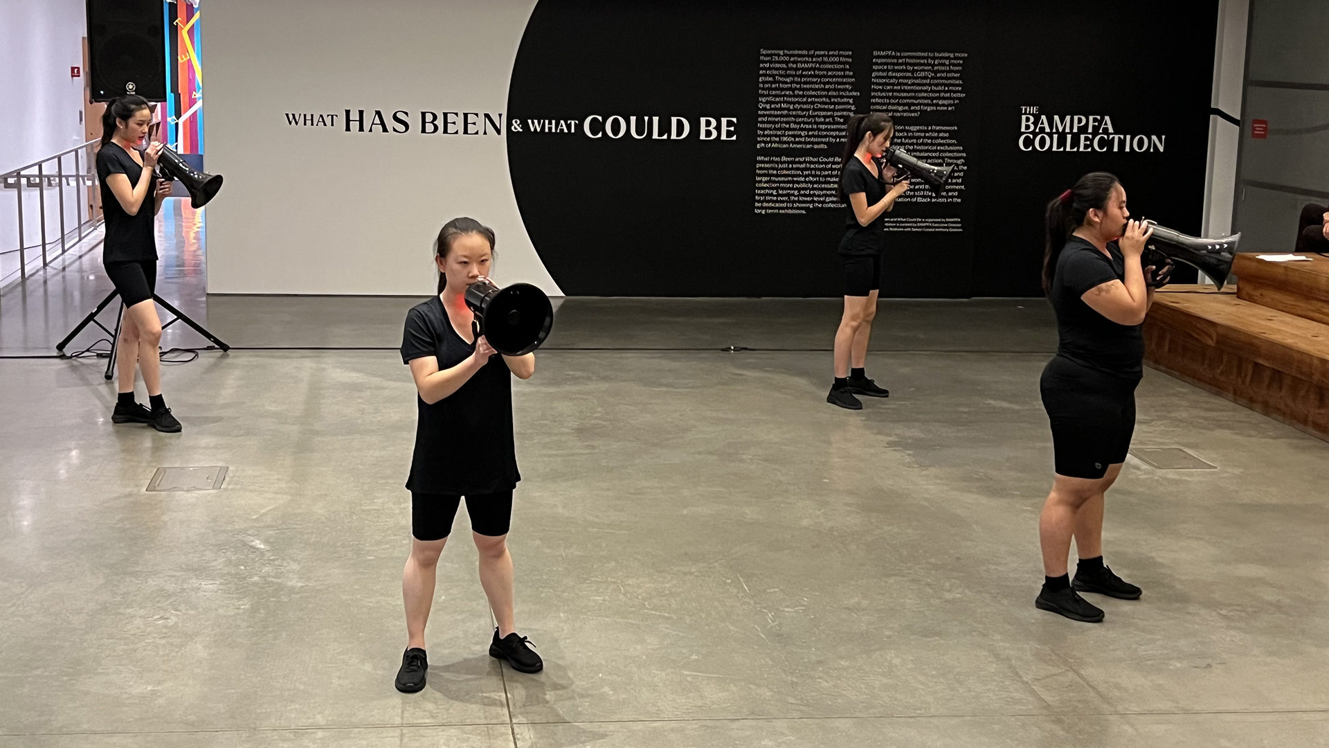 four dancers wearing all black hold bullhorns up to their mouths during a dance performance in a gallery space