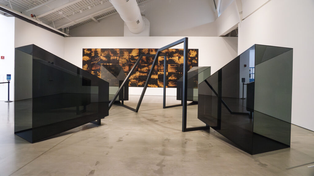 Two large, shiny black rectangular objects sit on the right and left, parallel to each other in a gallery. An artwork made of panels that look like they've been burned is on the center wall behind the objects. 