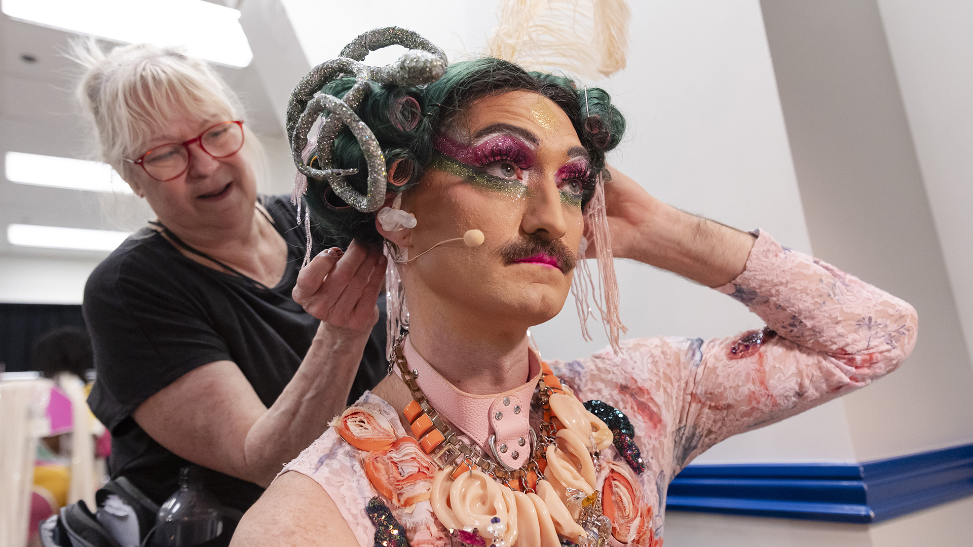 a wardrobe supervisor helps attach a wig to a performer with greenish hair in curlers and a silver sequin snake atop his head. The performer has a mustache and long fake lashes with green and magenta eye makeup and is wearing a pink lace top and a necklace of plastic ears.