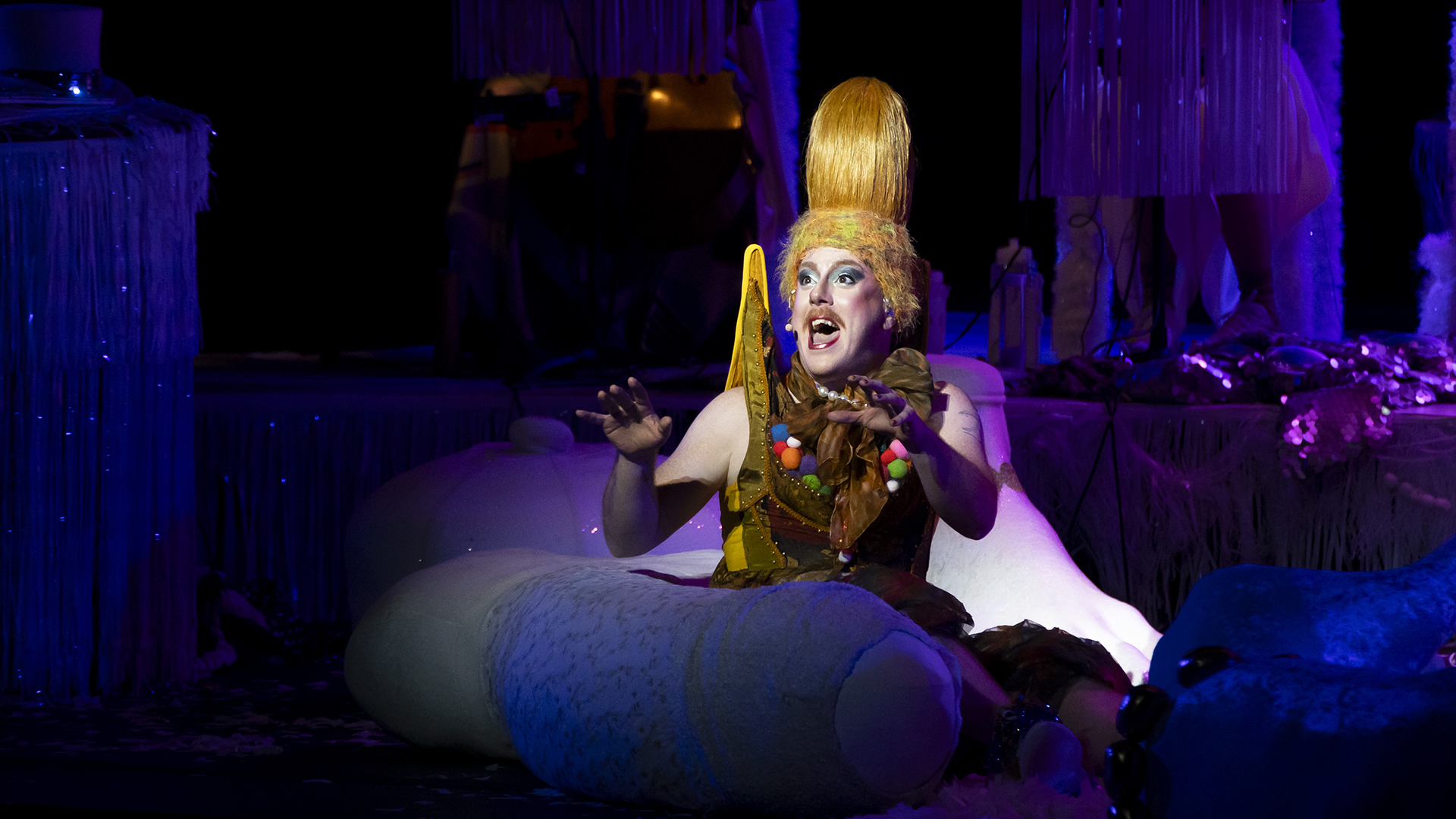 A man wearing a tall golden yellow wig in a kind of bun performs a solo in a spotlight on stage