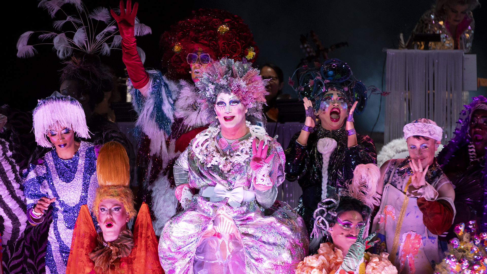 A half dozen people dressed in colorful, vibrant drag costumes perform a song on stage