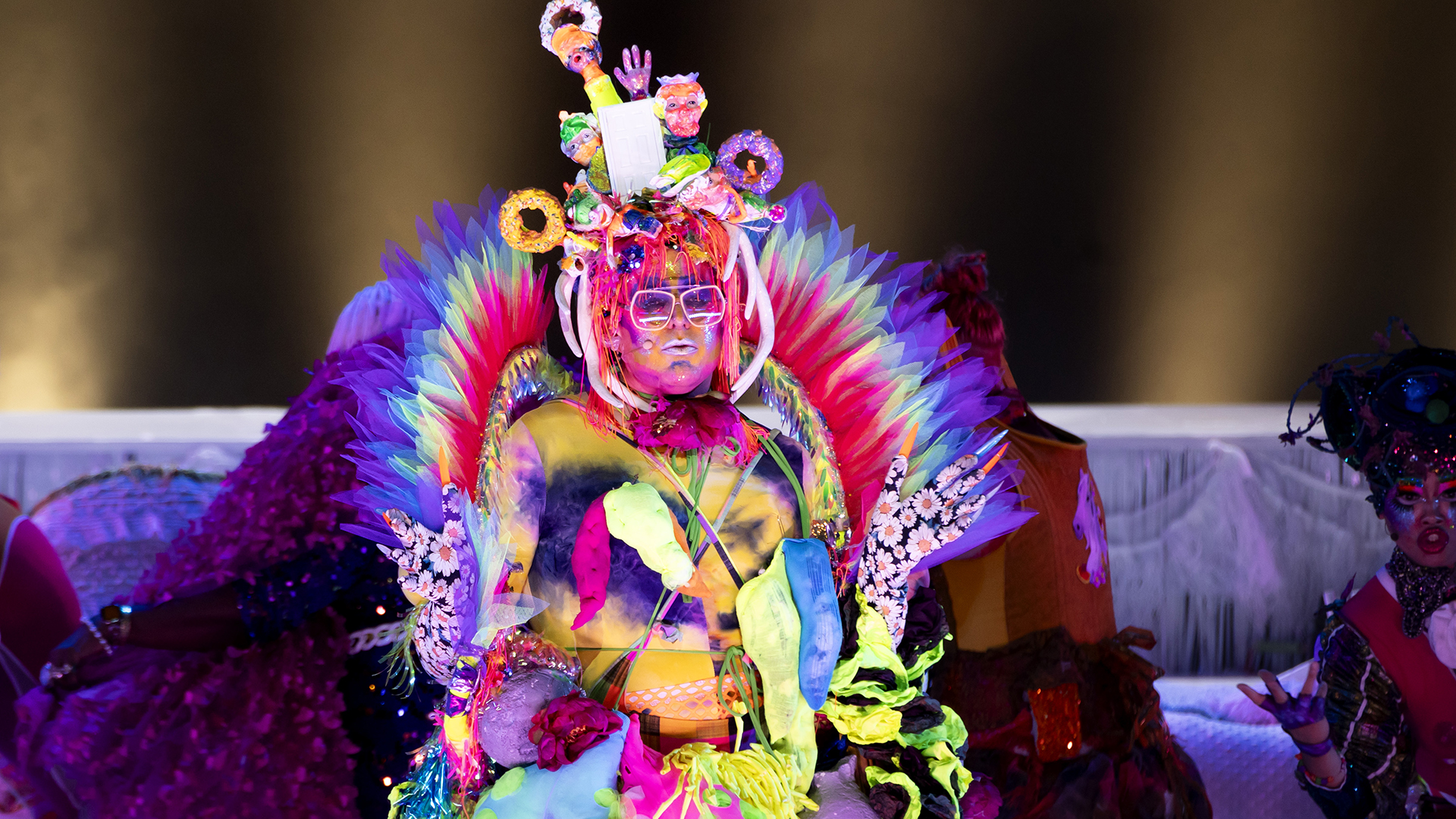 Machine Dazzle performs a musical solo on stage wearing bright neon rainbow feathers that go behind him and form a kind of halo around his shoulders and face. He is also wearing a neon outfit with long daisy-print gloves and headwear with little bejeweled ceramic heads, hands and donuts sticking out of it.