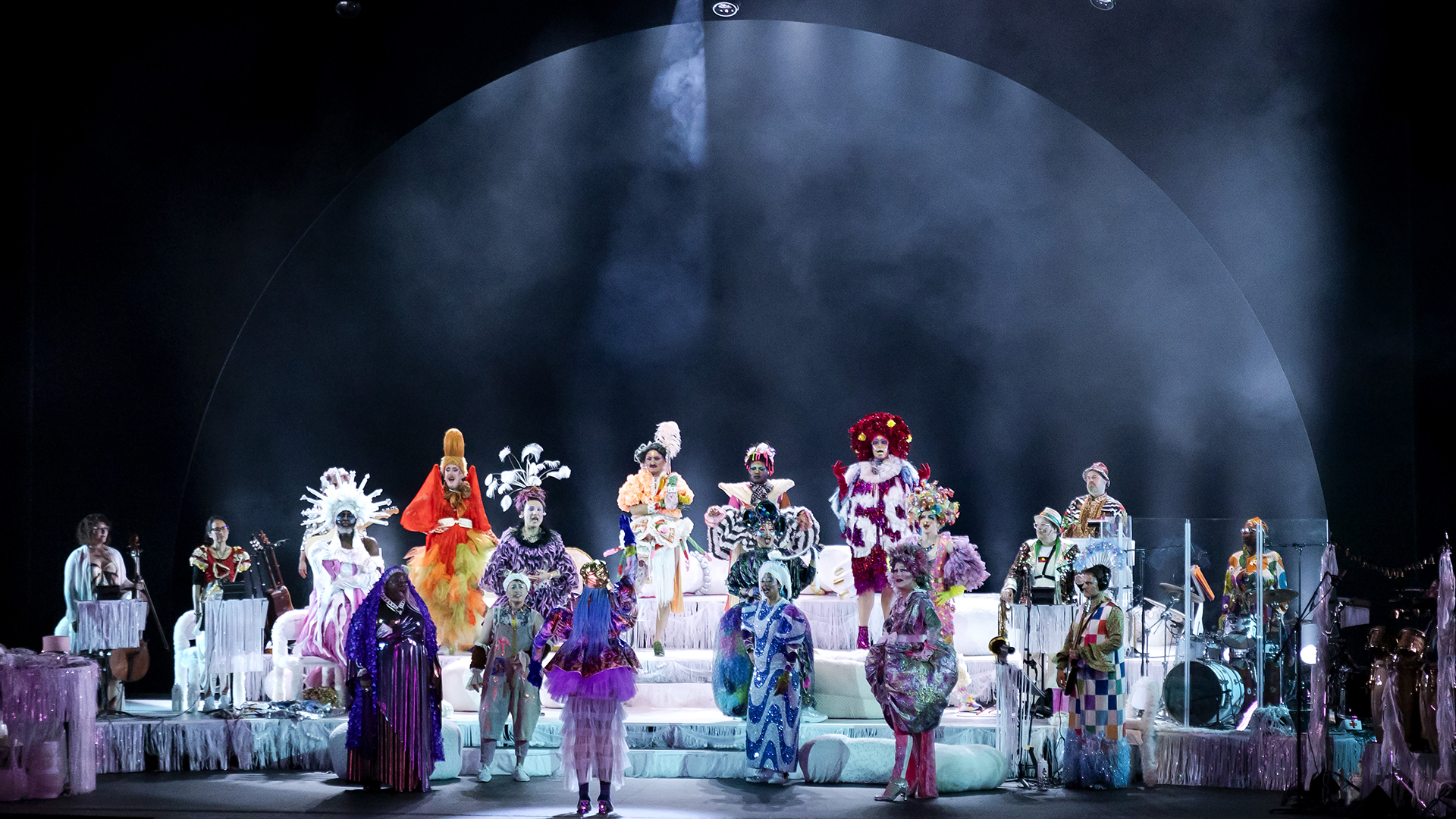 22 performers stand on stage, all dressed in vibrant costumes, sing a song together with a shape of a moon projected on the wall behind them