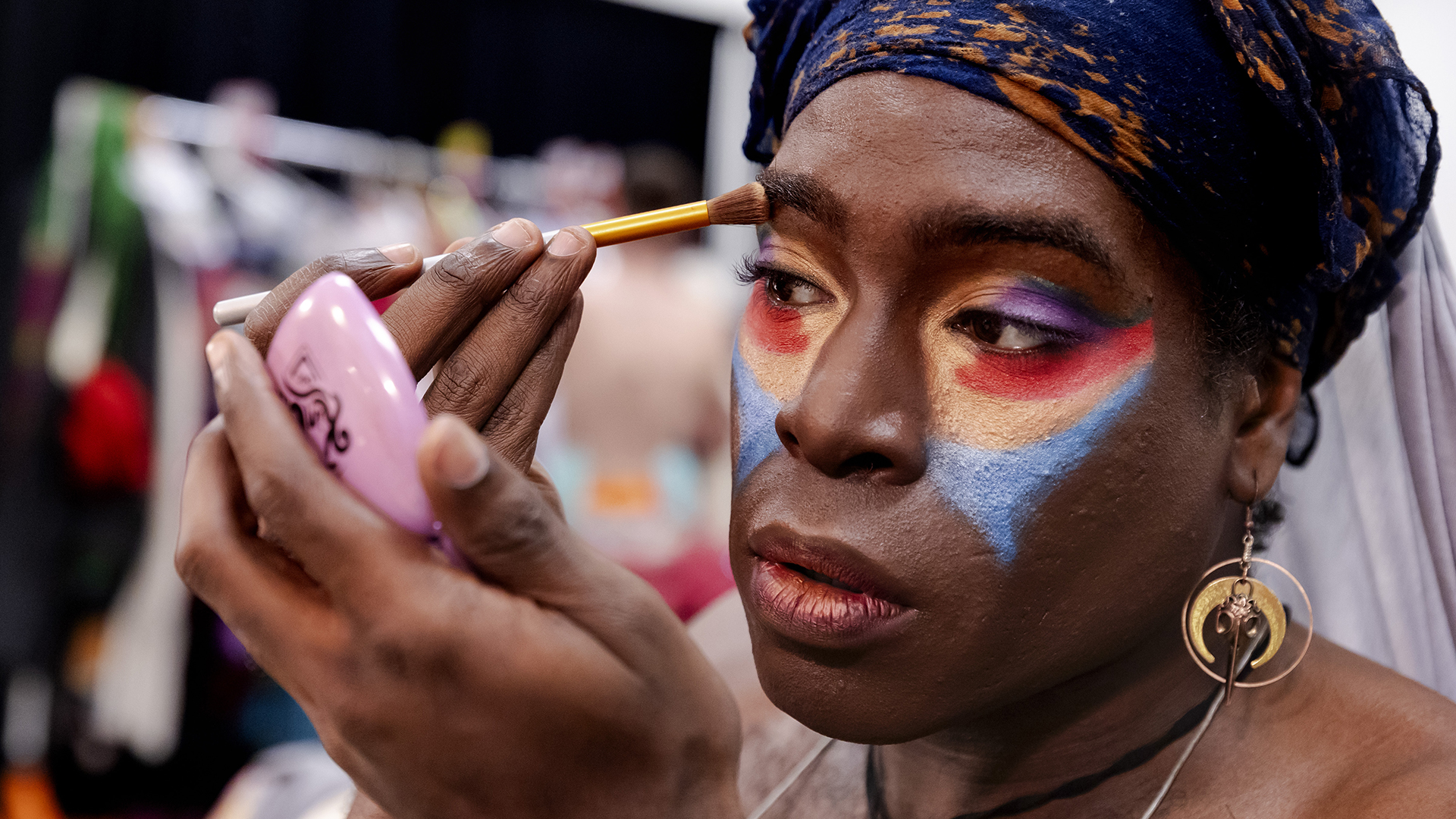 A closeup of a performer's face as they apply colorful makeup around their eyes while looking into a small compact mirror