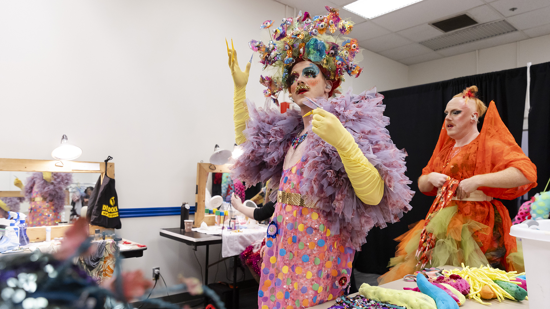 two performers get dressed in costumes in a dressing room — one person wearing a polkadot pink dress with a fluffy purple shrug made of tulle and headwear with fake eyeballs popping out of it and the other person in a bright orange top with a big colorful skirt