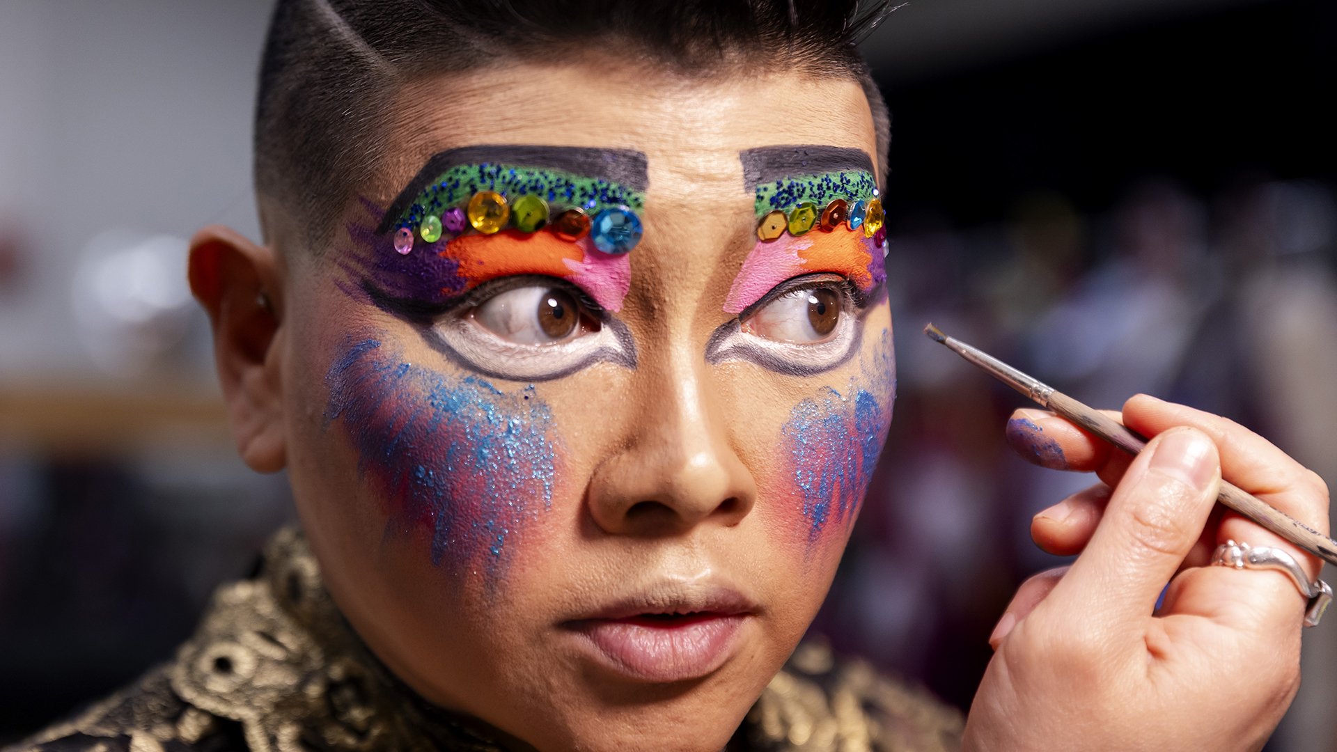 A closeup of a performer as they apply colorful makeup to their face