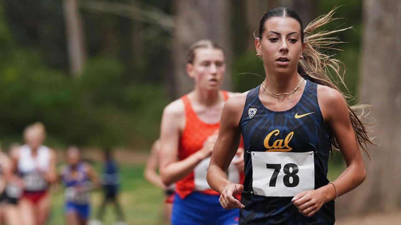 two women runners competing in an open field, against a forest backdrop