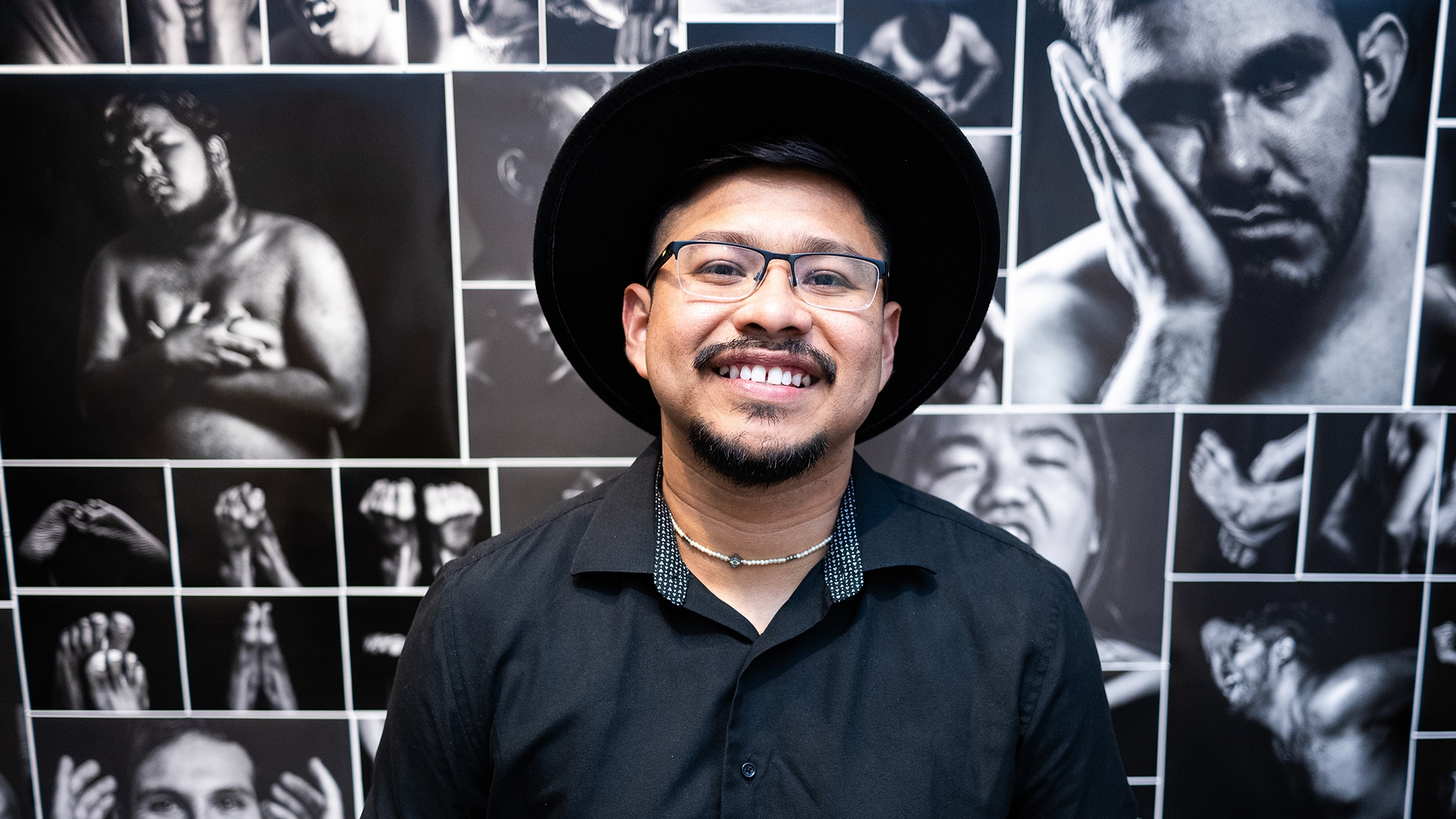 Brandon with a mustache and glasses and wearing a hat smiles and stands in front a wall of black-and-white photos of men is a range of poses that convey vulnerability