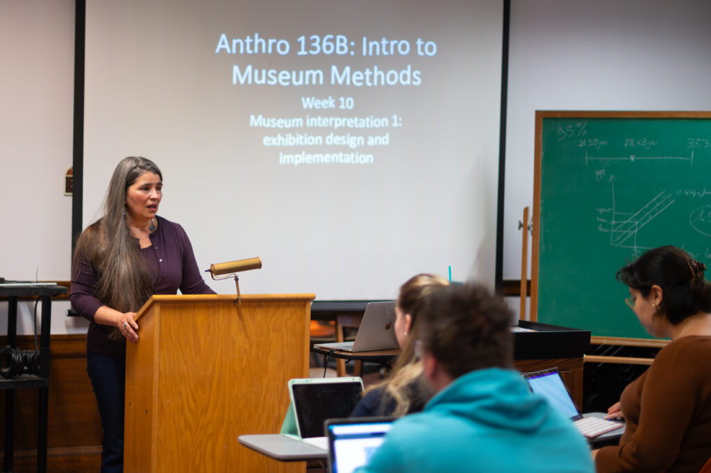 a photo of a person at speaking at a lectern to a class of students with a screen in the background about museum methods