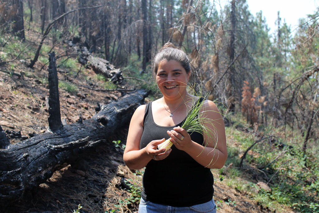 Carolyn wearing a tank top stands in a forest next to a black, burned tree holding grass and smiling