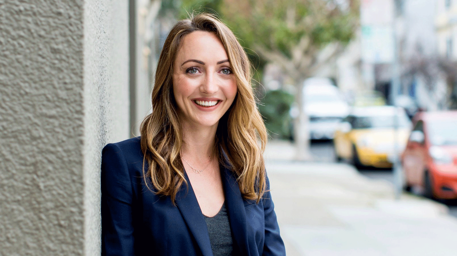 Informal portrait of Charlotte Hill, UC Berkeley political scientist, leaning against a building wall with an urban streetscape behind her