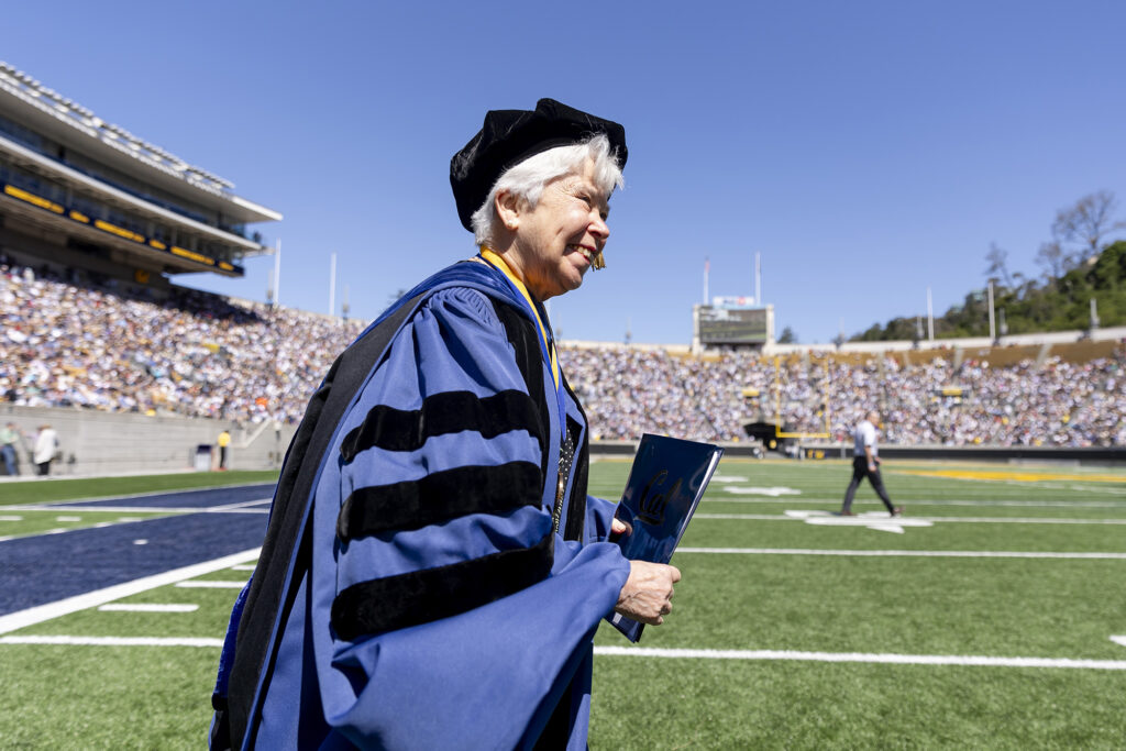 UC Berkeley Chancellor Carol T. Christ, wearing a commencement cap and blue robe, walking across a green football field.