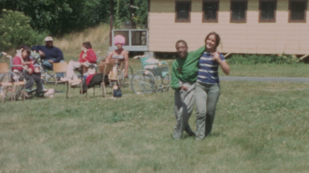 Two teenagers walk across a field while others in wheelchairs and chairs sit and relax in the background