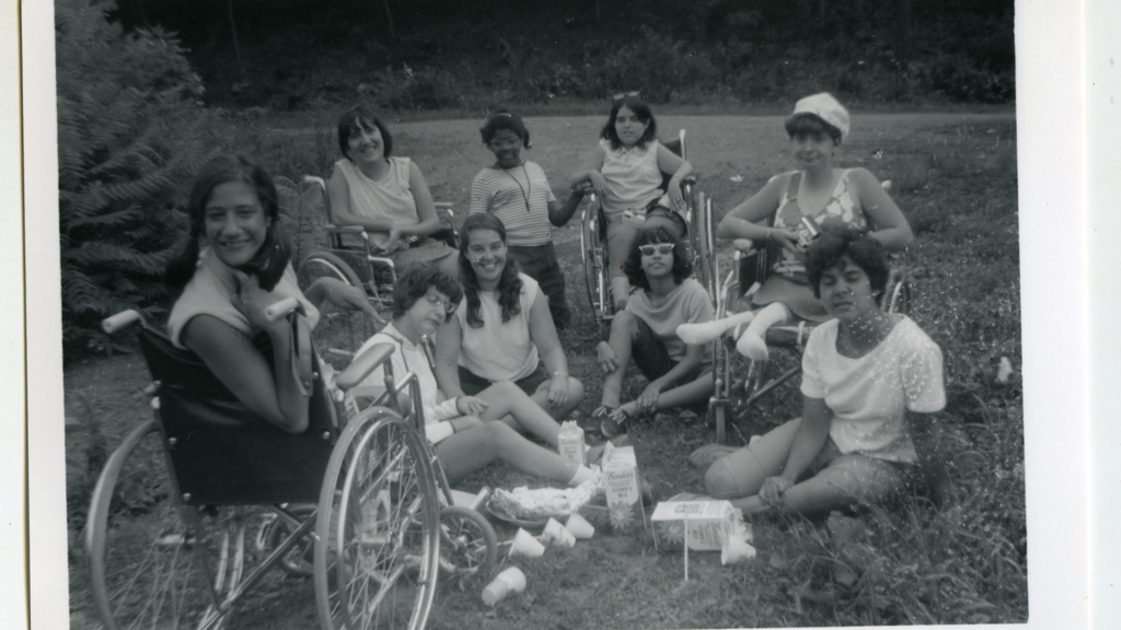 Black-and-white photo of nine people sitting on the grass and in wheelchairs in the summer at Camp Jened