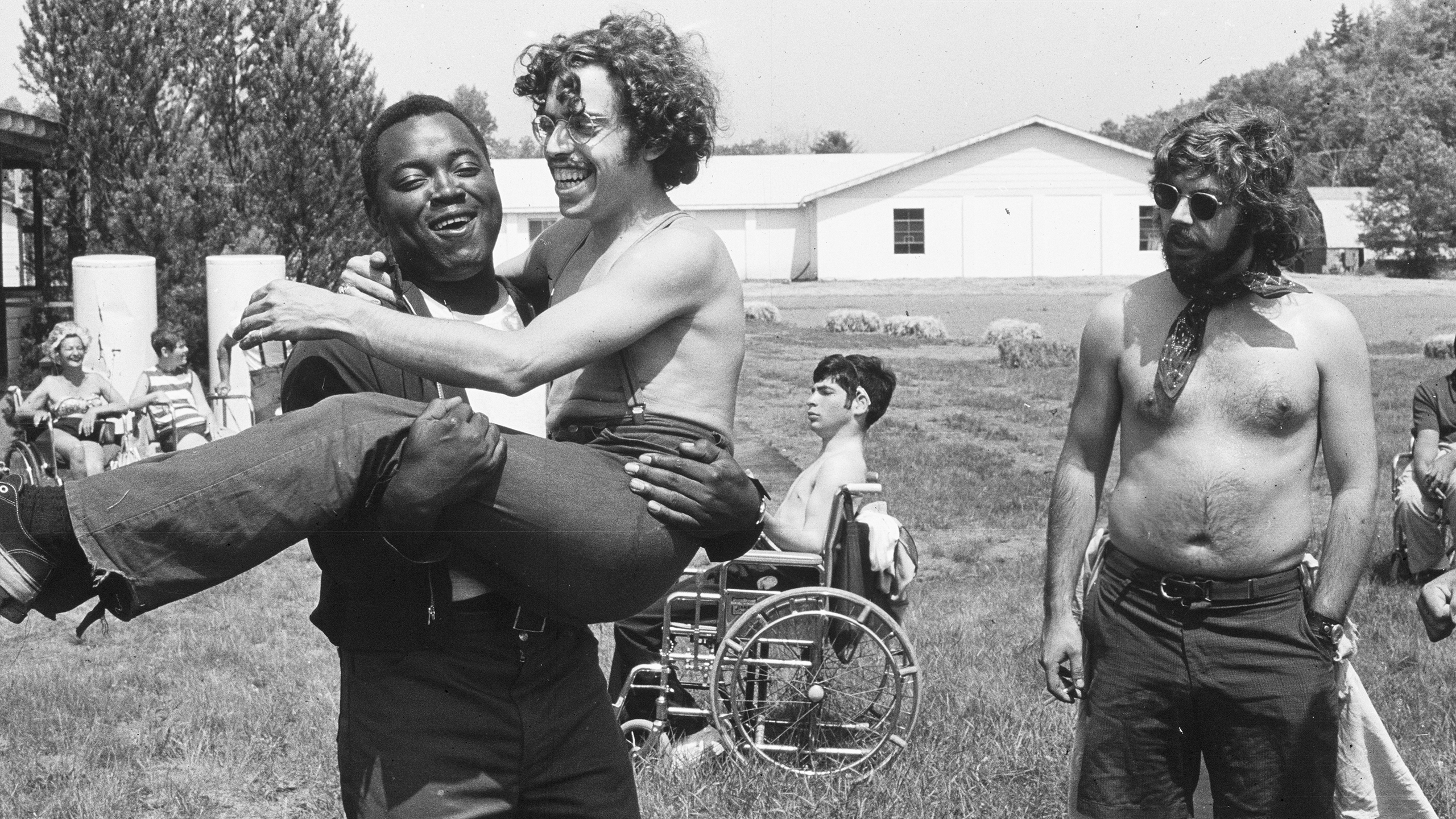 a black-and-white photo of people at Camp Jened, a summer camp for disabled people in the 1970s. A young man carries another young man while they laugh and as the shirtless director of the program looks on and campers in wheelchairs chat in the background.