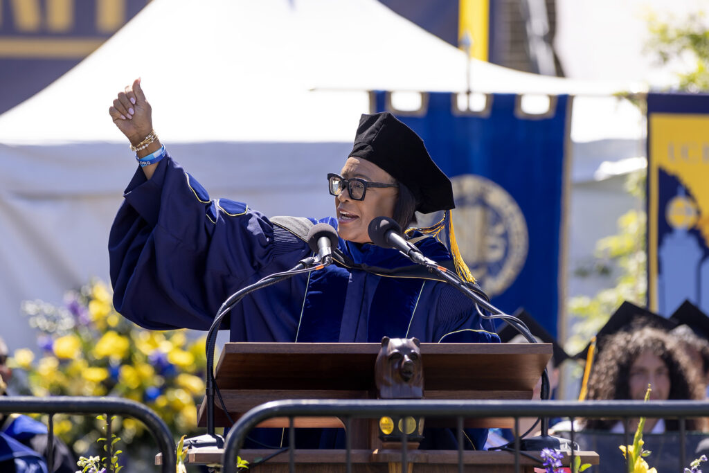 Commencement speaker Cynthia "Cynt" Marshall, wearing a cap and gown, gestures to the crowd.