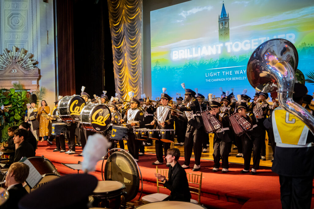 Members of the Cal Band perform on a large stage in the Oakland Scottish Rite Center, where a celebration was held Feb. 29, 2024, to celebrate the close of the Light the Way fundraising campaign. Large Cal drums are in the front of the band, as well as a tuba. The band is wearing crisp uniforms that include hats that each has an upright white feather on the top. Behind them is an image of the Campanile and the words 