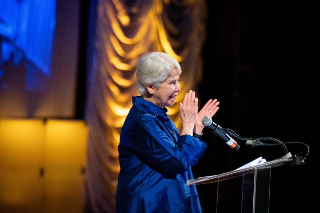 Chancellor Christ wears a bright blue jacket and applauds the success of the Light the Way fundraising campaign on a stage at the Oakland Scottish Rite Center. She is smiling and looking at the audience from a podium.