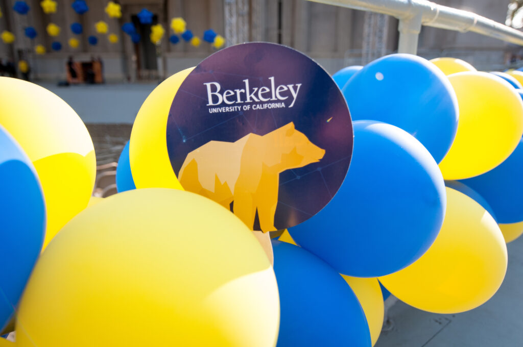 Atop a cluster of blue and gold balloons is a circular sign with a gold bear on it and the words UC Berkeley. The backdrop is the Greek Theatre.