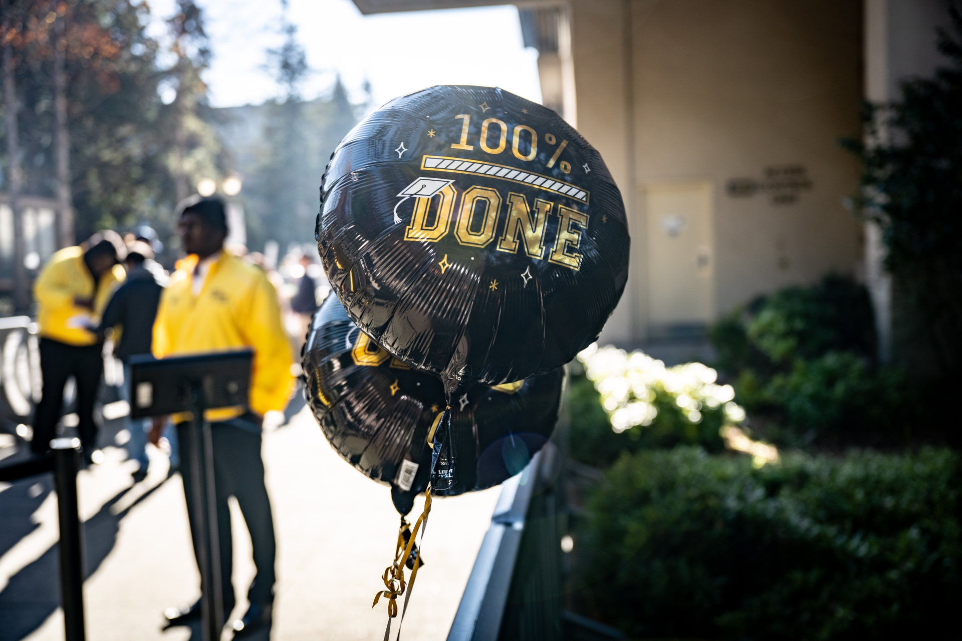 An image of a balloon with gold text that says "100% Done" in the foreground and people walking in the background.