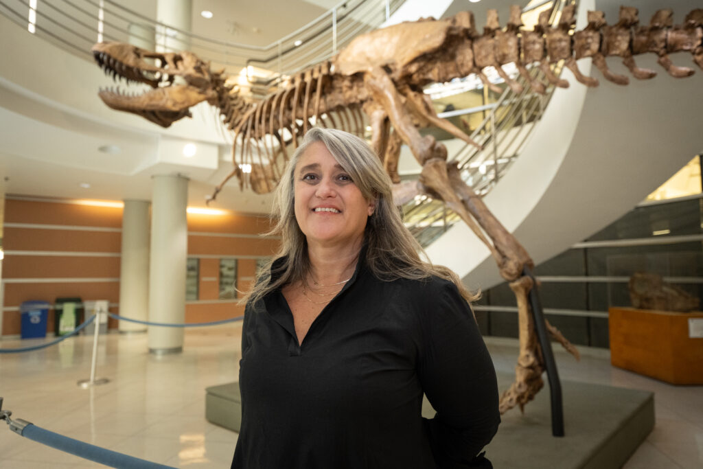 Gül Dölen with a black shirt and gray and black shoulder-length hair stands in in a building atrium in front of a skeleton of a T-Rex. 