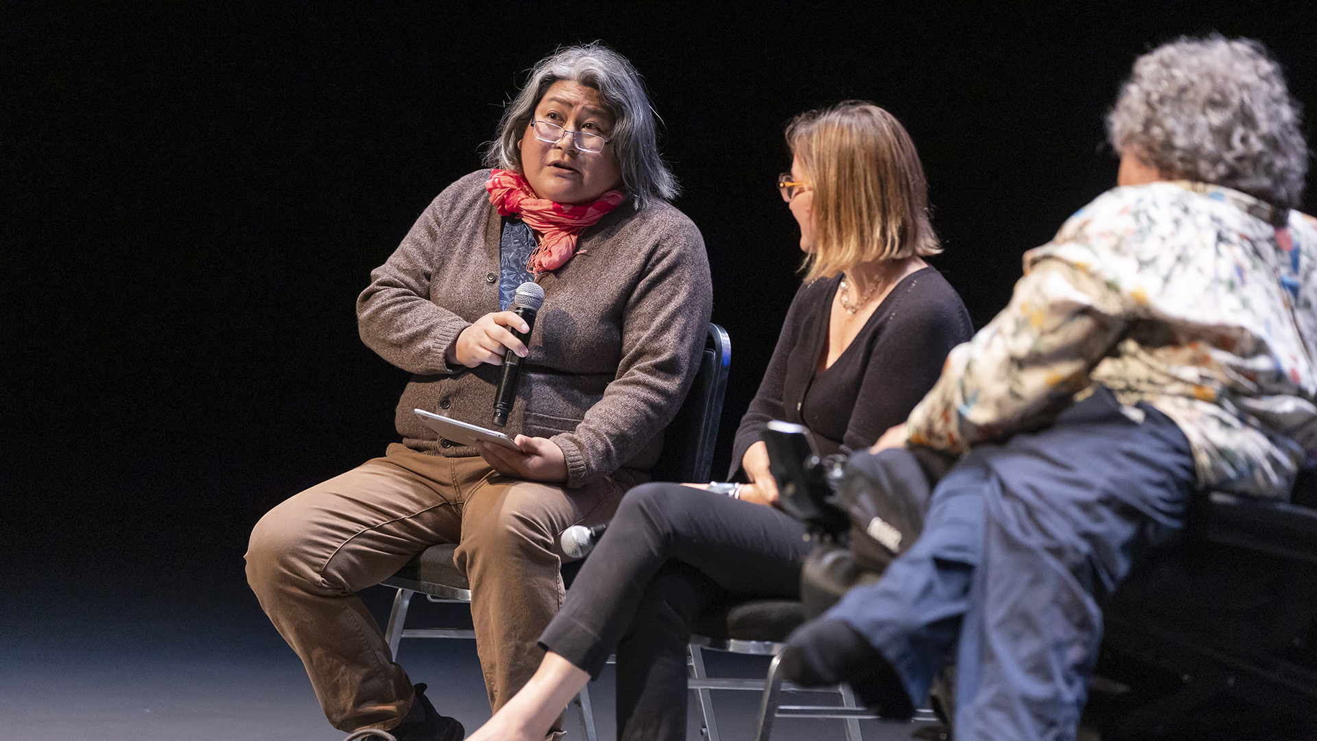 three people sit on stage, two with their backs to the camera and one person with shoulder-length salt and pepper hair and glasses facing forward holding a microphone. 