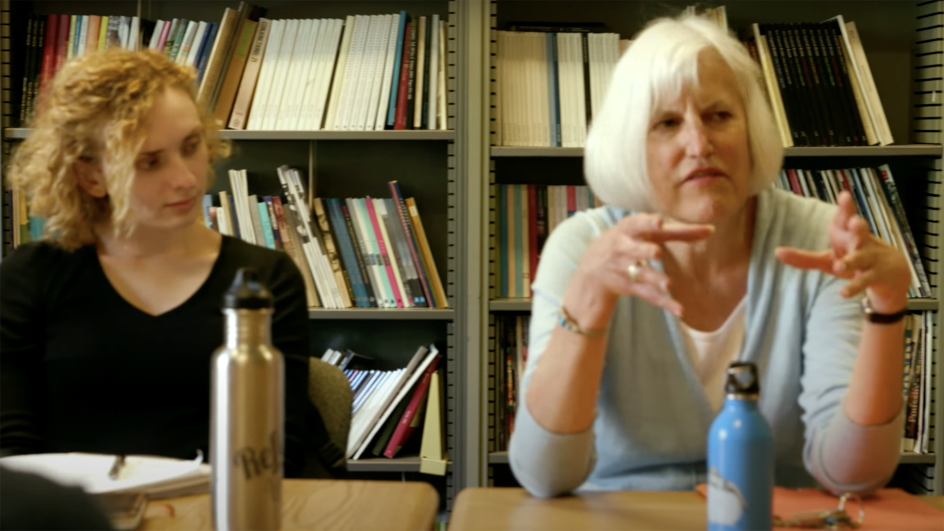 A woman with white, shoulder-length hair smiles and talks with two people in a room with a shelf of books in the background