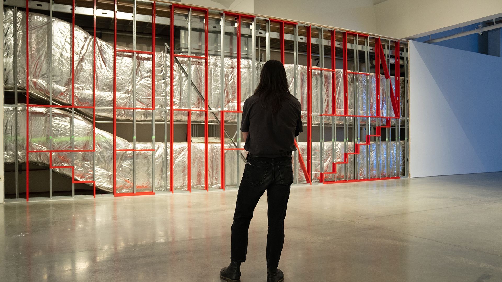 a person dressed in all black looks at a large artwork made of metal piping with designs painted on it in red